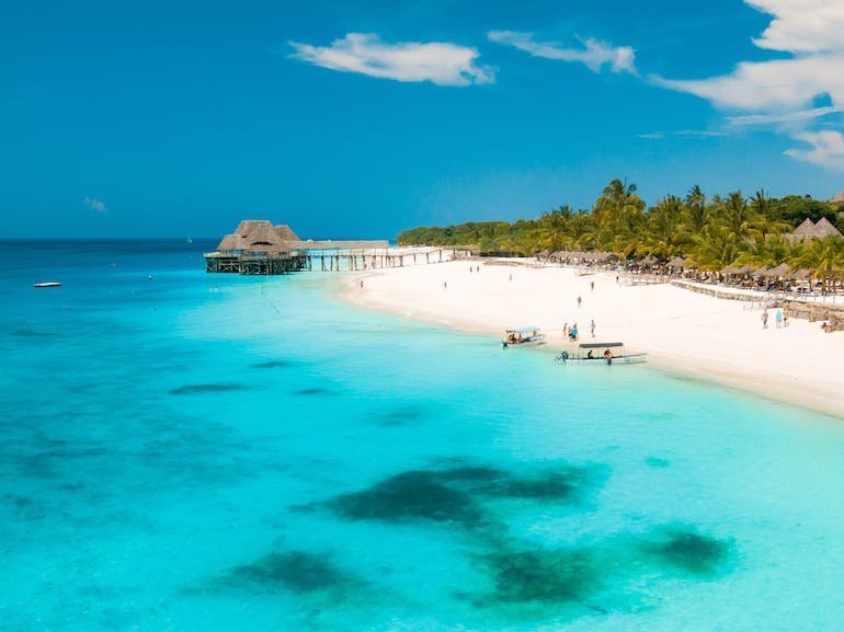 A lush forest meets the white sandy beaches in Zanzibar. Several people are seen on the beach and on boats with a pier in the distance. The turquoise waters shine brilliantly on this sunny day. 