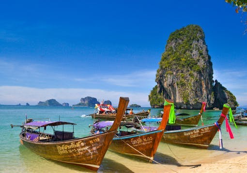 Traditional wooden boats in Thailand on the beach of Krabi are shown in shallow water with light blue water behind them and an impressive rock structure in the distance. Discover sustainable package trips in Thailand!
