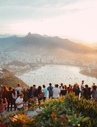 A line of people lean against a barrier admiring the view from a mountain top overlooking a city below with hills in the distance during sunset 