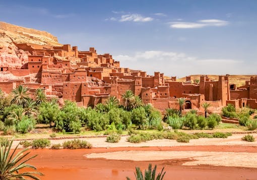 The historic orange village of Ait Benhaddou in morocco is shown from a near distance, with greenery in the front and a blue sky above.