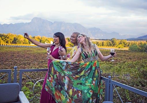 Three women with different skin tones wearing colorful dresses smile together while taking a selfie in the South African Cape Winelands, while holding glasses of wine.