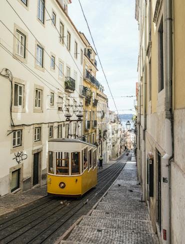 Yellow streetcar drives uphill surrounded by beige buildings in Lisbon, on a post guiding travelers about how to travel sustainably in Portugal.