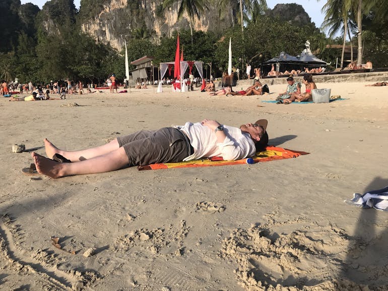 A White adult man lays on an orange and yellow beach towel in Thailand, wearing grey shorts, no shoes, a long sleeve shite shirt, watch, hat, and sunglasses. Other beachgoers are sittings and laying behind him. This is on a beach in Thailand on a sunny day. Greenery is in the background.