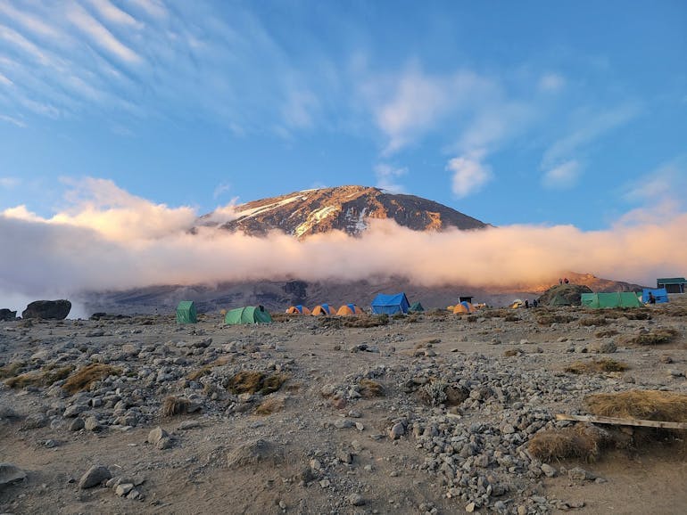 Mid climb on Mount Kilimanjaro in Tanzania, camp is set up with colorful blue and green and orange tents just below a layer of clouds, with the mountain peak behind and above the layer of clouds. Terrain is rocky and gravelly, sky is blue with some wispy clouds.