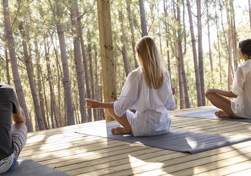 A Caucasian woman in a mid-length, white, long sleeve shirt dress sits in a meditative pose with her back to the camera. She sits on a yoga mat with a man sitting to her left and another Caucasian woman sitting to her right. They are on a slat wooden ground, overlooking thin trees with bright sunlight in the background.