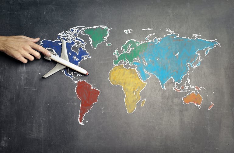 A colorful map where each continent is a different color is shown on a grey background, with a white hand pushing a small toy plane over a royal blue North America.