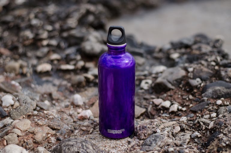 A reusable, stainless steel purple water bottle sits on a rock, with other rocks blurred in the background.