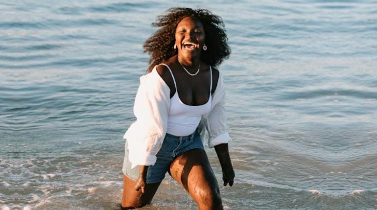 A young black woman with long curly coarse hair wearing jean shorts, a white tanktop, and a white open over shirt laughs joyously as she splashes through shallow water at sunset.