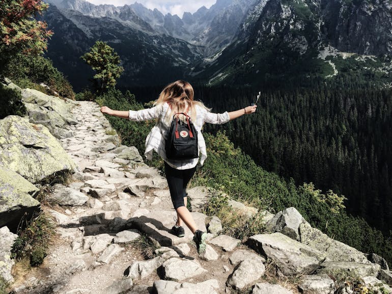 A young woman in black capris and a long sleeve open shirt skips along rocks on a hiking trail with her arms gleefully out to the side. She is in the mountains and there are peaks in the distance.