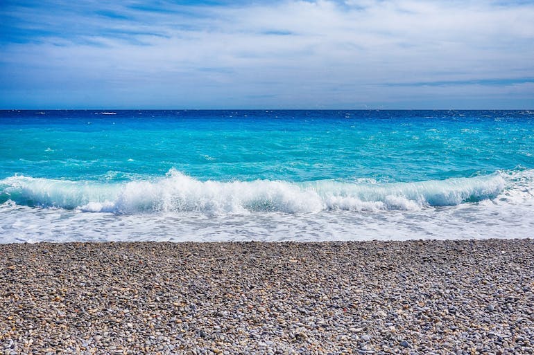 The purely azure blue waters are vibrant in the sunshine on the French Riviera. There is a pebble beach leading up to the lapping ocean and the sky has light clouds across it.