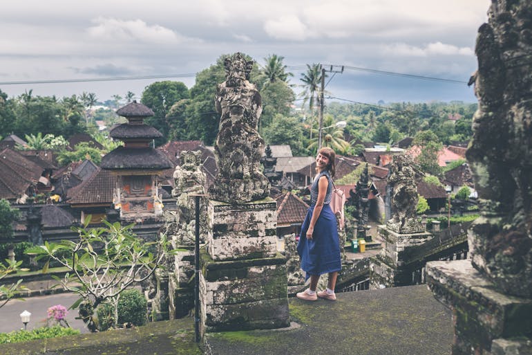 A female, White traveler wearing a blue dress smiles over her shoulder with a scenic view in the background. | Photo by Artem Beliaikin on Unsplash