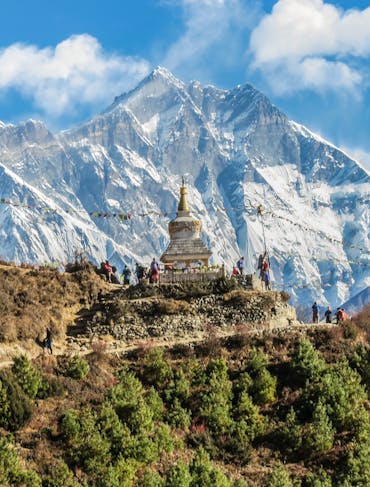 A stunning view of Mount Everest in Nepal is shown from across a green and brown forest, with a temple in the distance, looking small in front of the mighty mountain. The sky is bright blue with wispy clouds in the center and to the right of the mountain.