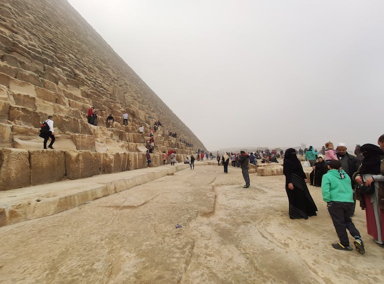 Overcrowding at the Pyramids of Giza in Egypt. Tourists are standing on the pyramid and to the right of it, milling about on a cloudy day.