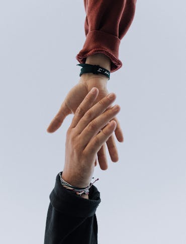 Two hands extend toward each other to grab one another, one from the bottom of the frame and the other from the top of the frame. Both are wearing dark long sleeves and some bracelets. The hands are white.
