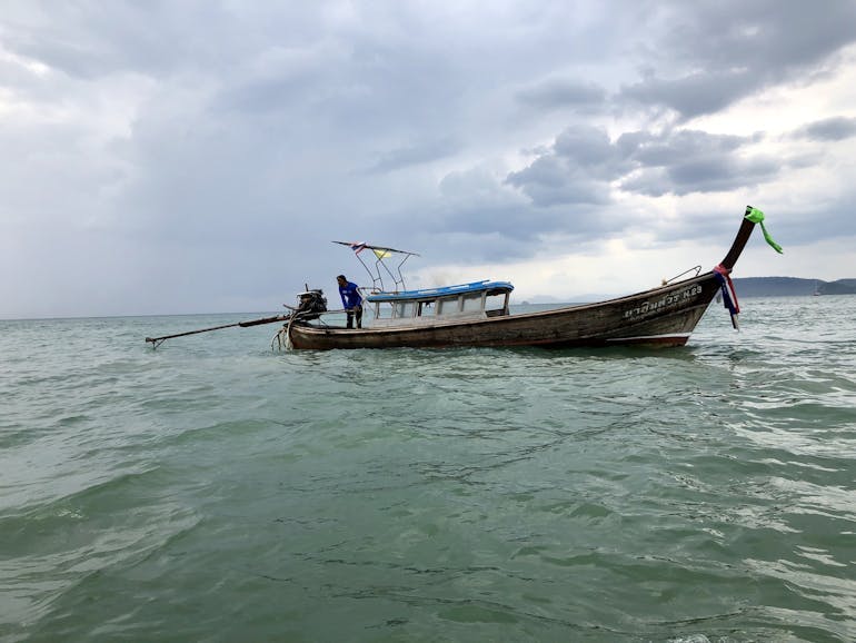 A small traditional wooden boat in Thailand is empty except for its captain, who gently steers the boat through the dark green water on a cloudy day