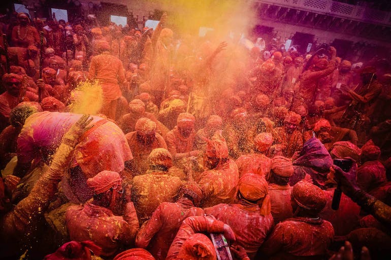 A large group of people covered in orange and yellow powder. One person to the left is in the midst of throwing the yellow powder on to the group
