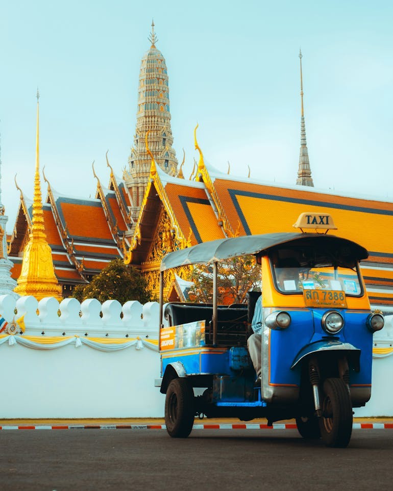 A blue and yellow tuktuk in Bangkok, Thailand is shown in from of a bright yellow and orange temple.