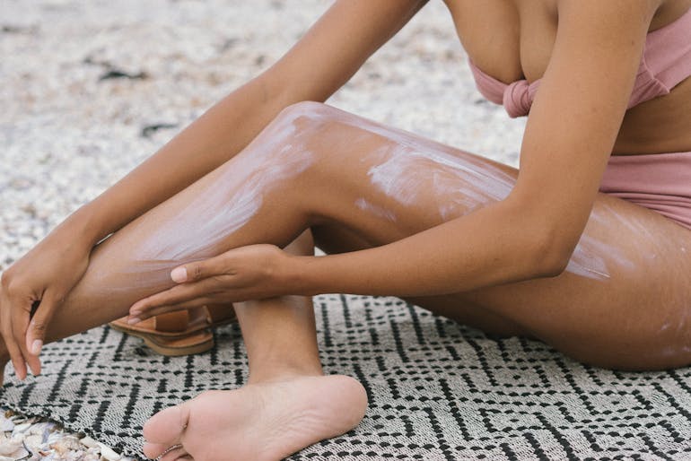 A tan woman applies white sunscreen to her legs while sitting on a patterned town at the beach in a light pink two piece swim suit.