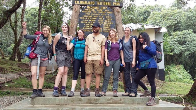 A group of 6 Caucasian women stand, 3 on each side of a Black man (the guide) in front of the Machame Gate wooden signpost with yellow lettering. This is just before they start the Machame Route to climb Mt. Kilimanjaro in Tanzania.