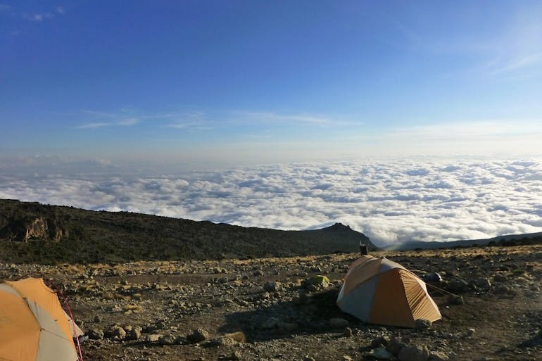 Two tents are shown on Mount Kilimanjaro in Tanzania overlooking a blanket of clouds down below, with blue sky above.