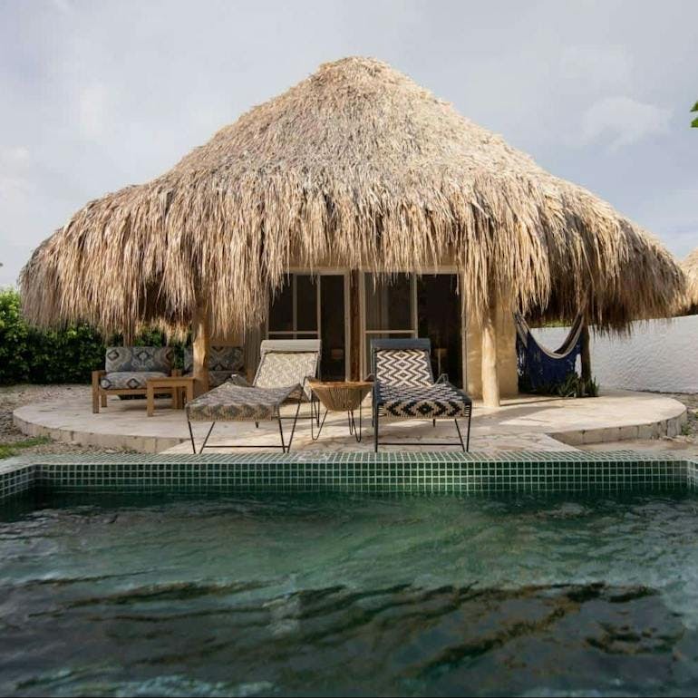 A bungalo over the water with a thatched room and two sunchairs in front on either side of a small table. A small couch is in the background.
