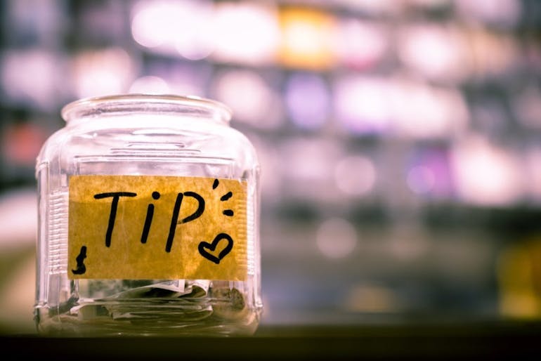 A glass jar with a brown paper taped to it that says "Tip" with a heart drawn is shown in focus, the rest of the image is blurred. | Photo by Sam Dan Truong on Unsplash