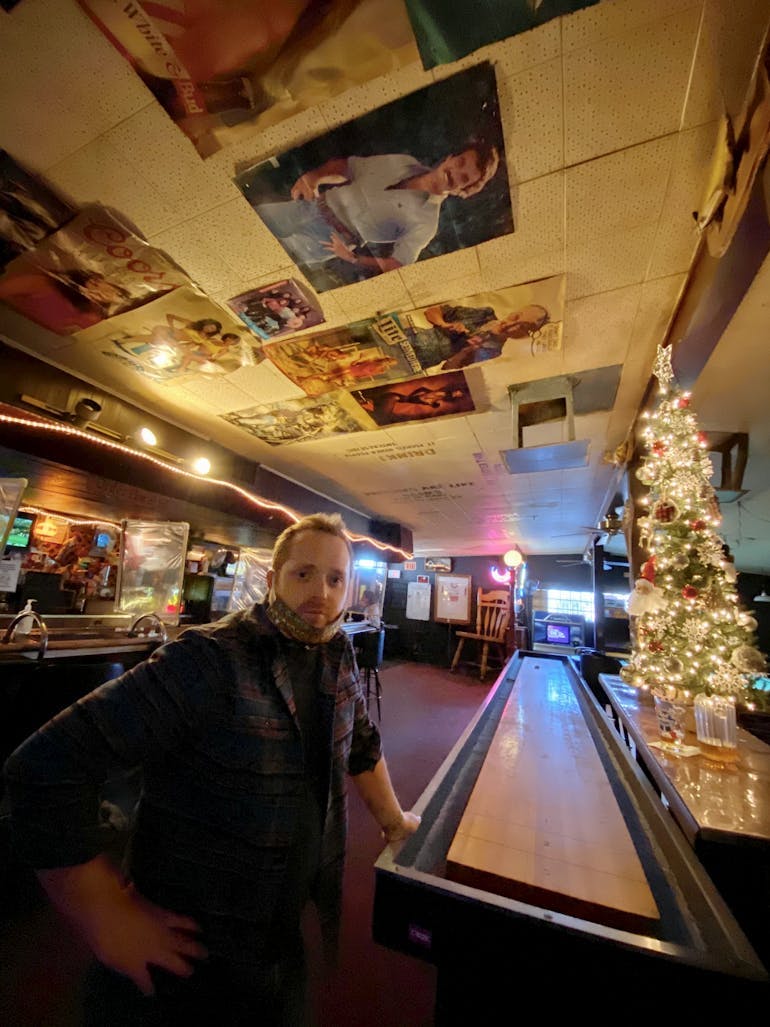 A White adult man with short ginger hair and a goatee, wearing a mask around his chin and in a long-sleeved plaid shirt stands in a dimly lit bar in Egypt. A Christmas tree is lit to the right, a shuffleboard game that the man in leaning on, and old posters are on the ceiling.