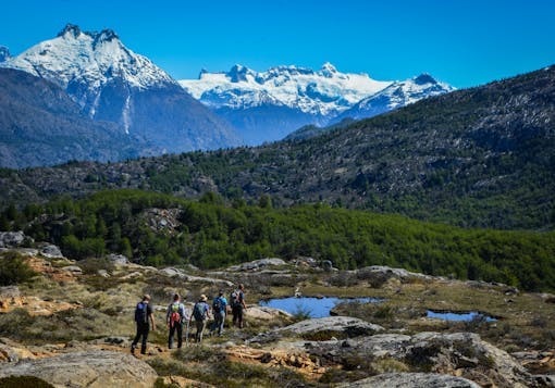 A group of 5 trekkers walk along the Gaucho Way in wild Chilean Patagonia on a responsible trekking tour. They walk over mixed terrain of dirt and rocks, with a small pond nearby and snow-capped mountains in the distance.