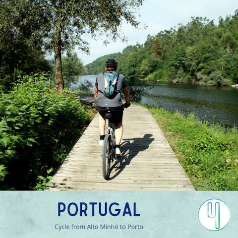 An adult Caucasian man bikes along a boardwalk with lush greenery to his left and a flowing river to his right in Porto's Alto Minho region.