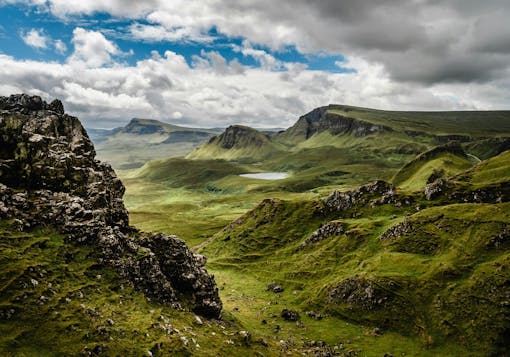 The Isle of Skye is shown in Scotland with mossy, craggy green hills and a cloudy sky with some blue peeking through.