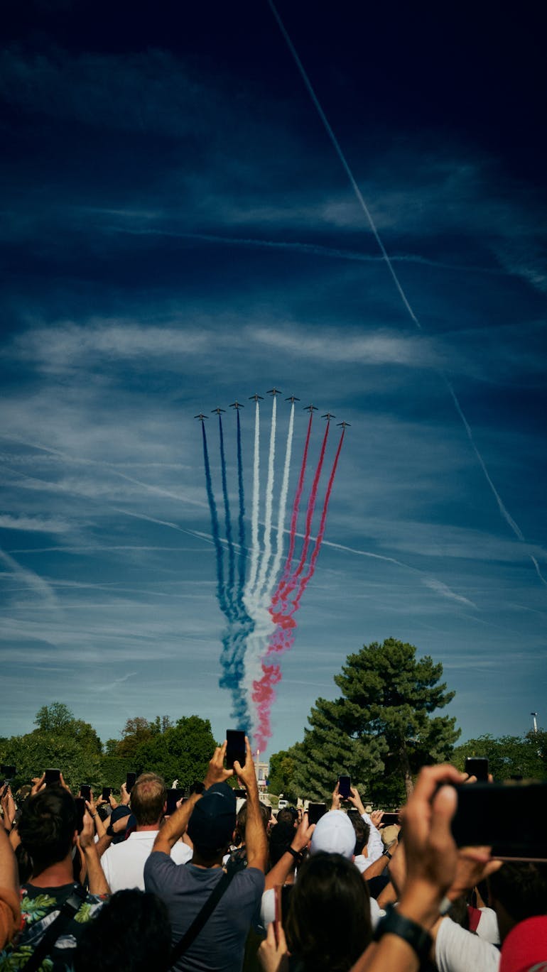In the clear sky you can see 9 planes flying in formation with the French national colours trailing behind them. Together they make the famous tri-colour flag of France. The plane are flying above a crowd of people looking up to the sky admiring the show, most of which have phones in their hands. 