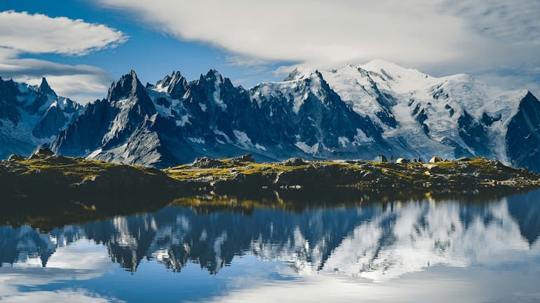 A sharp, jagged mountain range spans the image with some grassy areas resting at the foot of the mountain. In front of the green space, there is a body of water where you can see the mountain's reflection. 