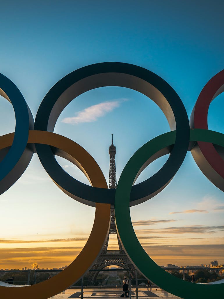 The Olympic Rings are in the foreground and fill most of the image with the Eiffel Tower peaking through in the background during sunset. 