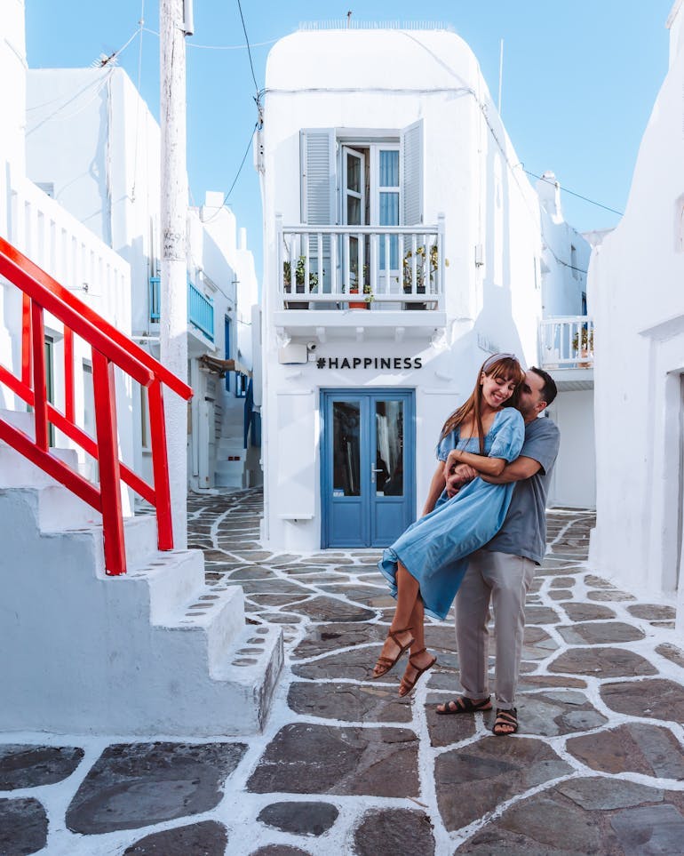 A young heterosexual couple stand on stone flooring in Greece, the man lifting up the woman from behind, with the classic white washed buildings in the background and a red stairway railing to the left, and blue doors behind them.
