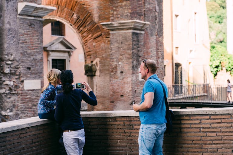 A group of three young adult friends stand around in a foreign country admiring the nearby architecture and taking a picture of it with one's digital camera.