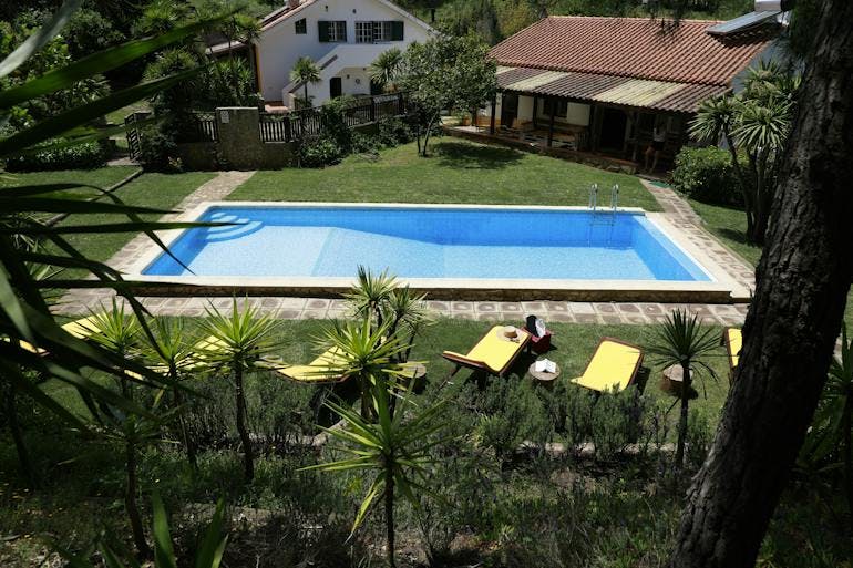 A sparkling blue pool sits in the sunshine with yellow sun chairs in the foreground. There is green grass all around and some of the quaint accommodations surrounding it.