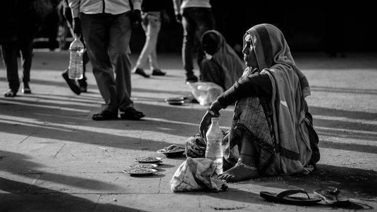 A black and white photo of an Indian woman sitting on the street with her sandals to the side, a bottle of water in front of her, and dishes in front of her with things - perhaps herbs - on front of them. Unclear if she is begging or selling something.