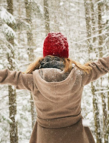A woman with long, straight blond hair wearing a beige coat, red had, and black gloves stands with her back to the frame with her arms outstretched, forming a T, amidst a snowy forest of thin trees.