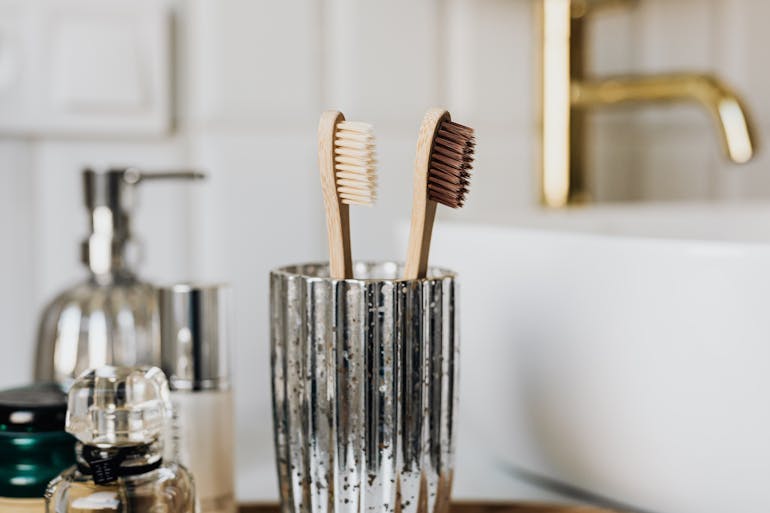 Two brown bamboo toothbrushes are in a glass cup by a sink in a bathroom, with other bathroom products blurred in the background.