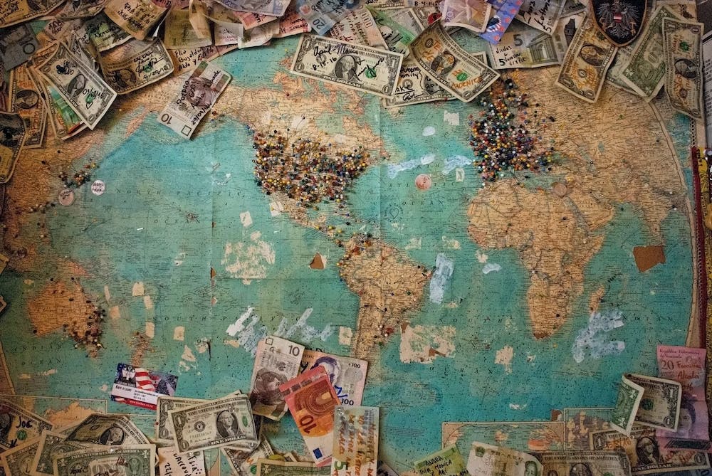 An image of a map with money of different currencies laying around its borders and pins pushed into many spots around the world, particularly in North America and Europe. | Photo by Christine Roy on Unsplash