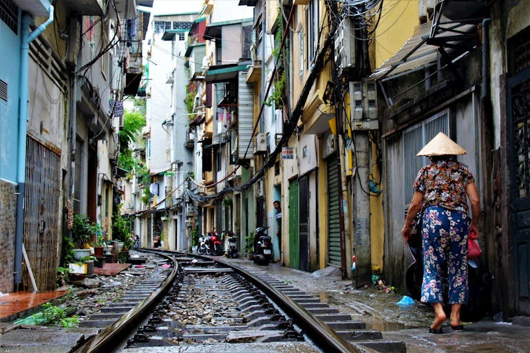 A woman in blue floral pants and darker floral short sleeve shirt and a traditional Vietnamese sunhat walks to the right side of a railroad traffic through a narrow street full of multi-level colorful houses.