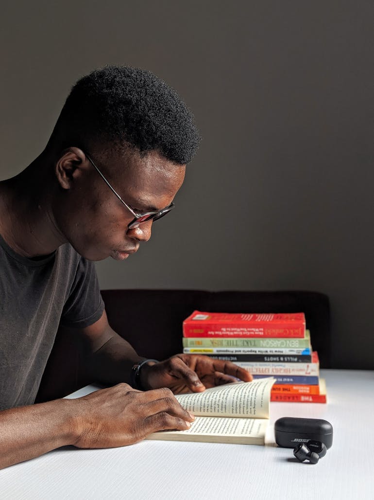 A young Black man sits and reads a book; we can see his side profile. A stack of several books are in the background and there is a grey wall behind him.