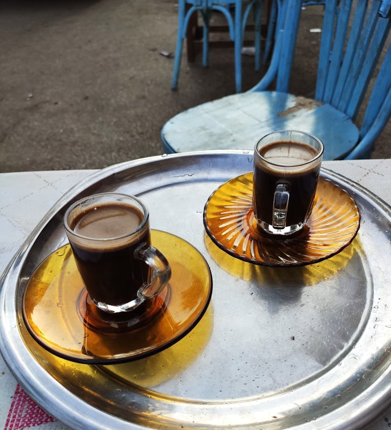 Two glass coffee cups filled with coffee site on yellow glass plates on top of a circular aluminum tray, resting on a table top with blue chairs in the background. Coffee is in in Egypt.