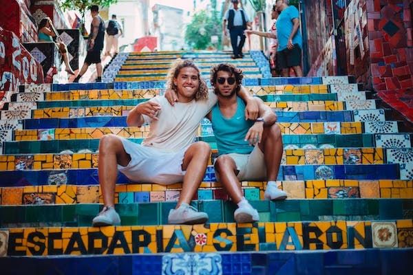 Two male, White friends in casual shorts and t shirts sit with their arms around each other on colorful steps, smiling at the camera. | Photo by Elizeu Dias on Unsplash