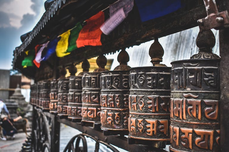 Brown metallic Buddhist prayer wheels are shown in Nepal in a long row, with colorful prayer wheels hung above them.