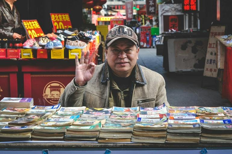 An Asian man sits in a brown jacket and baseball cap making the "A-OK" symbol with his right hand and smiling at the camera as he sits behind a stack fo books and magazines laid out at a market. OTher shops are selling shoes and other merchandise behind him.