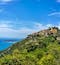 The village of Eze sits perched high atop a hill covered in lush greenery. The sparkling blue Cote d'Azur is to the left. The sky is blue. 