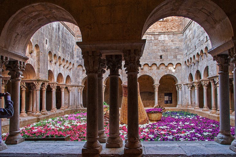 The annual Flower Festival in Girona can offer views like this, of bright pink, purple, and white flowers filling an open-air courtyard. The building surrounding the courtyard has archways and columns forming a square around it. 